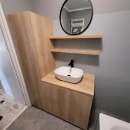 Nowoczesna łazienka z drewnianą szafką pod umywalką nablatową, okrągłym lustrem z czarną ramą i dwiema p&oacute;łkami na szarych płytkach. W tle widoczny grzejnik drabinkowy odbijający się w lustrze.