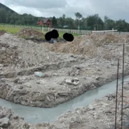 Zalane betonem wykopy pod fundamenty domu jednorodzinnego, widoczne zbrojenia, hałdy ziemi, w tle zabudowa mieszkalna i zalesione wzgórze.