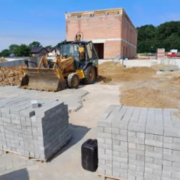 Żółta koparko-ładowarka Case 580 Super LE na placu budowy z paletami szarych kostek brukowych i fundamentem budynku w tle, w słoneczny dzień.