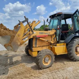 Żółta koparko-ładowarka Case 580 Super LE z łyżką pełną ziemi na placu budowy, widoczne fundamenty w tle, błękitne niebo z chmurami.