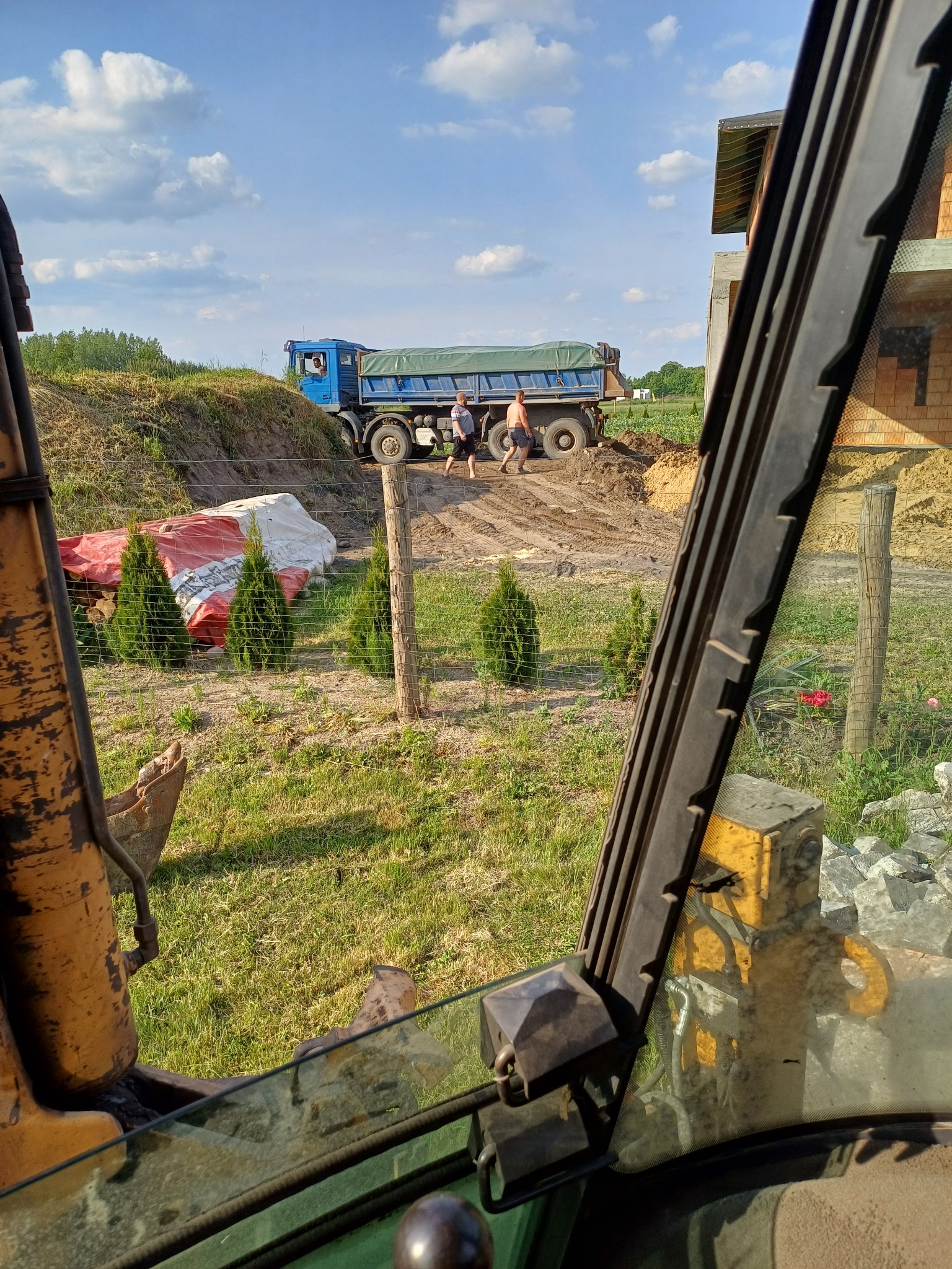 Widok z kabiny koparki na wywrotkę z zieloną plandeką na wzniesieniu, dwóch mężczyzn obok, w tle budynek w trakcie budowy, częściowo zasłonięty przez ramę okna koparki.