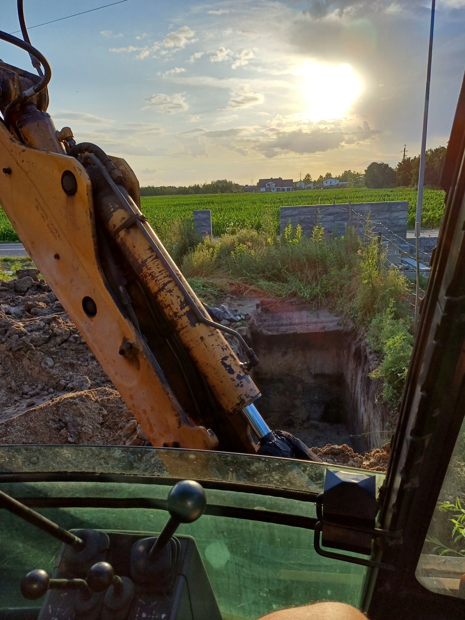 Widok z kabiny koparki na wykop w ziemi, z widoczną ścianą wykopu i fragmentem ramienia koparki na tle pola i zachodzącego słońca.