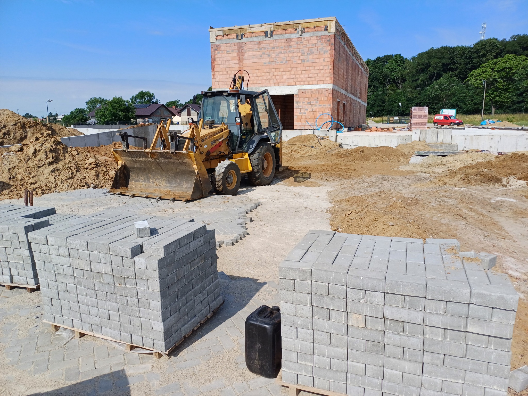 Żółta koparko-ładowarka Case 580 Super LE na placu budowy z paletami szarych kostek brukowych i fundamentem budynku w tle, w słoneczny dzień.