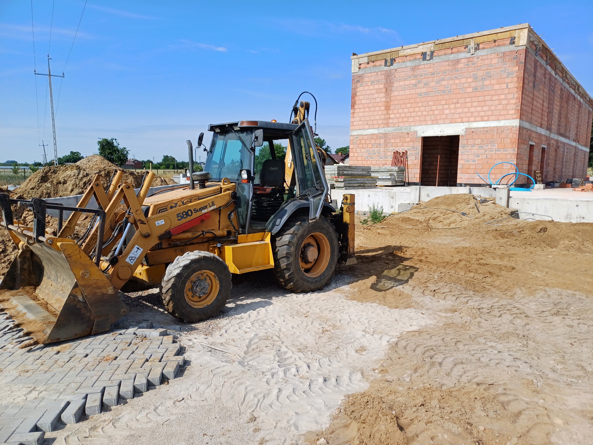 Żółta koparko-ładowarka Case 580 Super LE na placu budowy z częściowo ułożoną kostką brukową i fundamentem budynku z czerwonej cegły w tle.