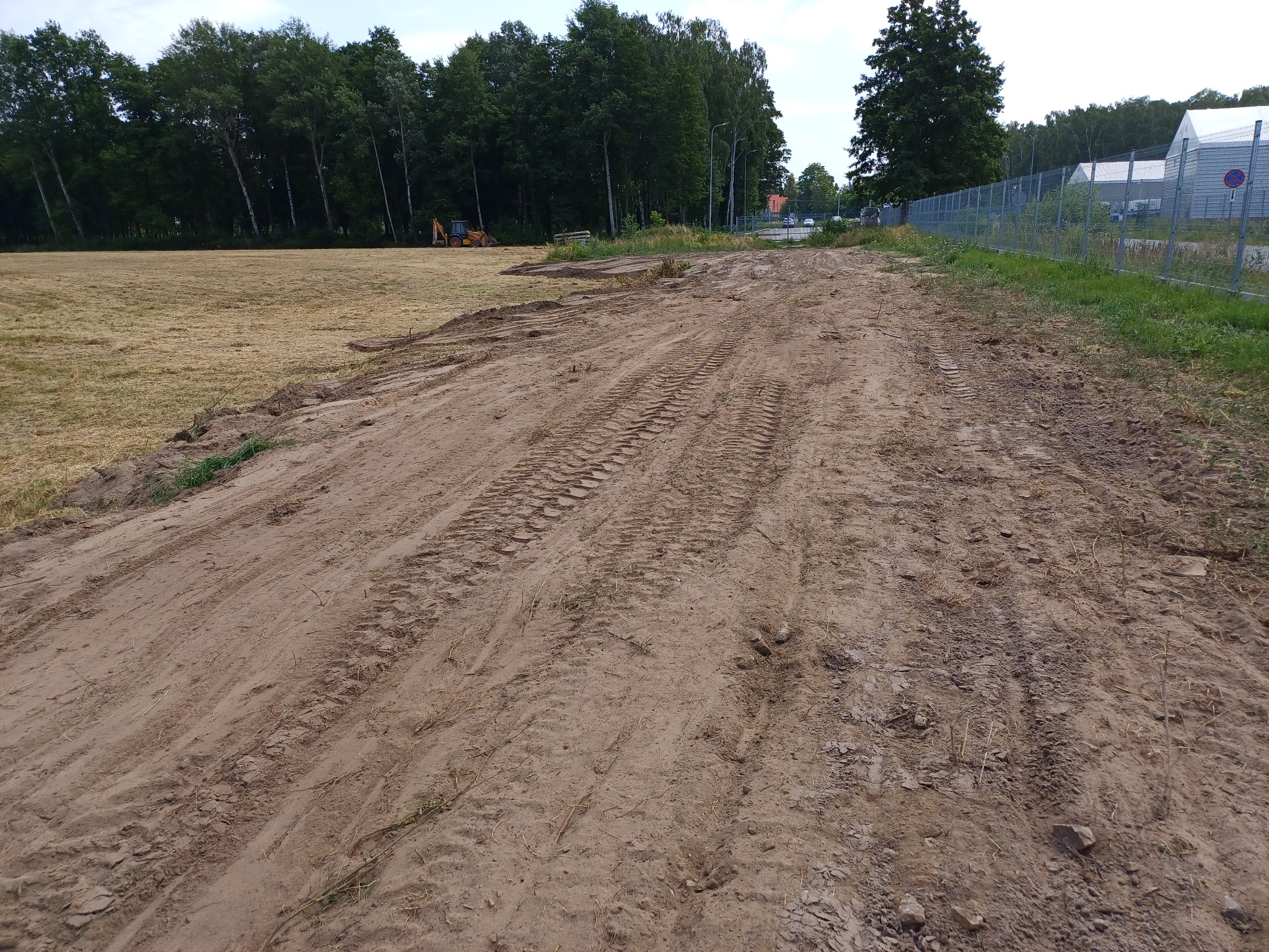 Ujeżdżona, piaszczysta droga z widocznymi śladami opon ciężkiego sprzętu, obok przygotowane pole i las w tle, z daleka widoczna koparka.