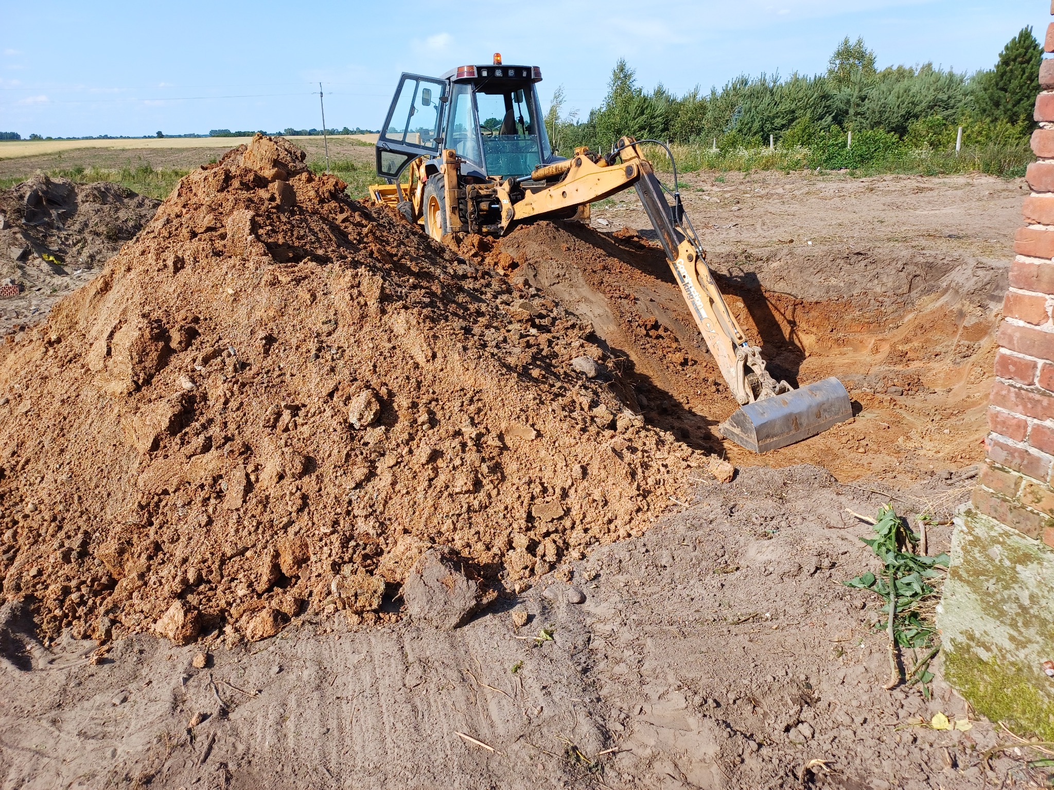 Żółta koparko-ładowarka podczas wykopywania fundamentów pod budynek, z widoczną stertą ziemi i fragmentem ceglanego muru.