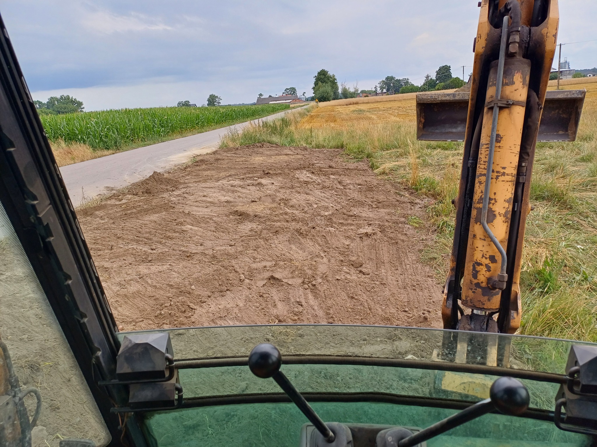 Widok z kabiny koparki na wyrównany teren pod drogę gruntową przy polu kukurydzy, z widocznym fragmentem ramienia koparki i elementami sterowania w kabinie.