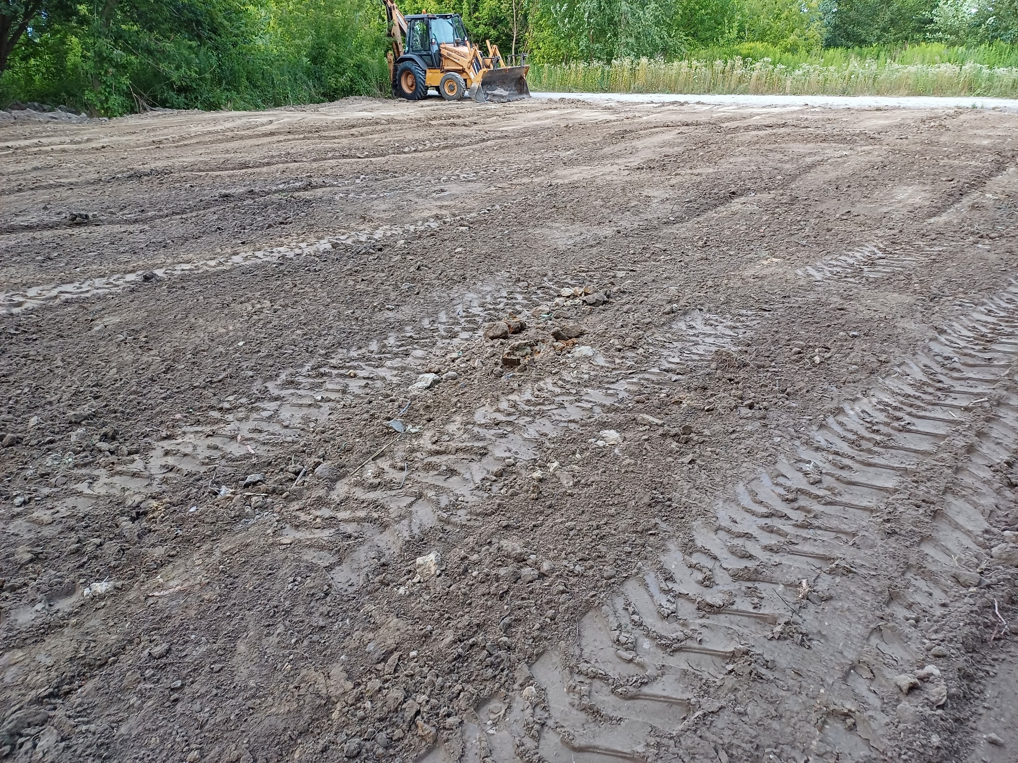 Żółta koparko-ładowarka na nierównym, błotnistym terenie z wyraźnymi śladami opon, z zielenią w tle.