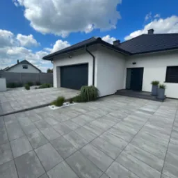 Nowoczesny podjazd z szarych kostek brukowych prowadzący do białego domu z czarną bramą garażową i dachem, ozdobiony trawami i białymi kamykami.