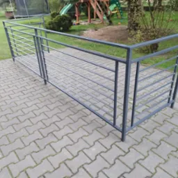 Szara metalowa balustrada o minimalistycznym designie, z poziomymi prętami, ustawiona na kostce brukowej w ogrodzie z placem zabaw w tle.