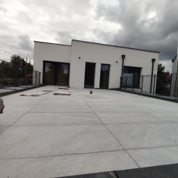 Jerzy Nowak - Jasnoszara, płytkowa powierzchnia tarasu na dachu nowoczesnego, białego budynku w trakcie prac wykończeniowych, z widocznymi elementami balustrady i fragmentem opakowania ochronnego.