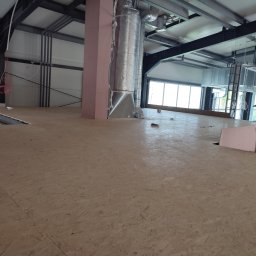 Jerzy Nowak - Wnętrze hali w trakcie budowy, widoczne płyty OSB tworzące podłogę, stalowa konstrukcja dachu, izolowane rury wentylacyjne i okna w tle.