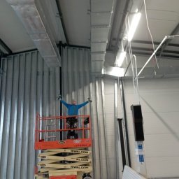 Jerzy Nowak - Pracownik na podnośniku nożycowym z uniesionymi rękami, trzymający wiertarki, widoczny na tle metalowej konstrukcji i instalacji wentylacyjnej w hali przemysłowej podczas montażu płyt karton gips.