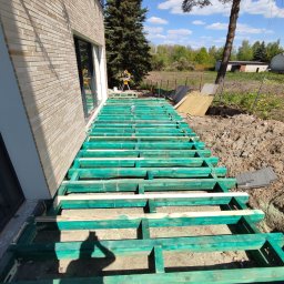 Jerzy Nowak - Konstrukcja nośna z impregnowanego drewna pod przyszły taras, widok z góry na szkielet, cień osoby fotografującej w lewym dolnym rogu, dom z cegły w tle, narzędzia na trawie.