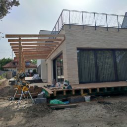 Jerzy Nowak - Budowa tarasu przy nowoczesnym domu z drewnianą konstrukcją pergoli i widoczną deską tarasową, narzędzia pracy w tle.