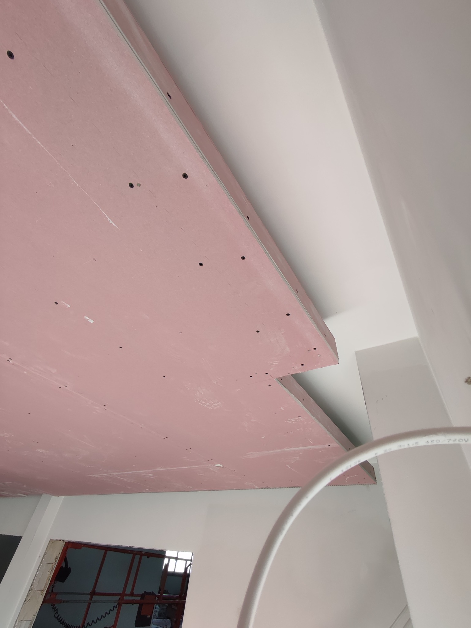 Montaż różowej płyty gipsowo-kartonowej na suficie, widoczne czarne śruby i białe ściany w tle, instalacja elektryczna częściowo widoczna.