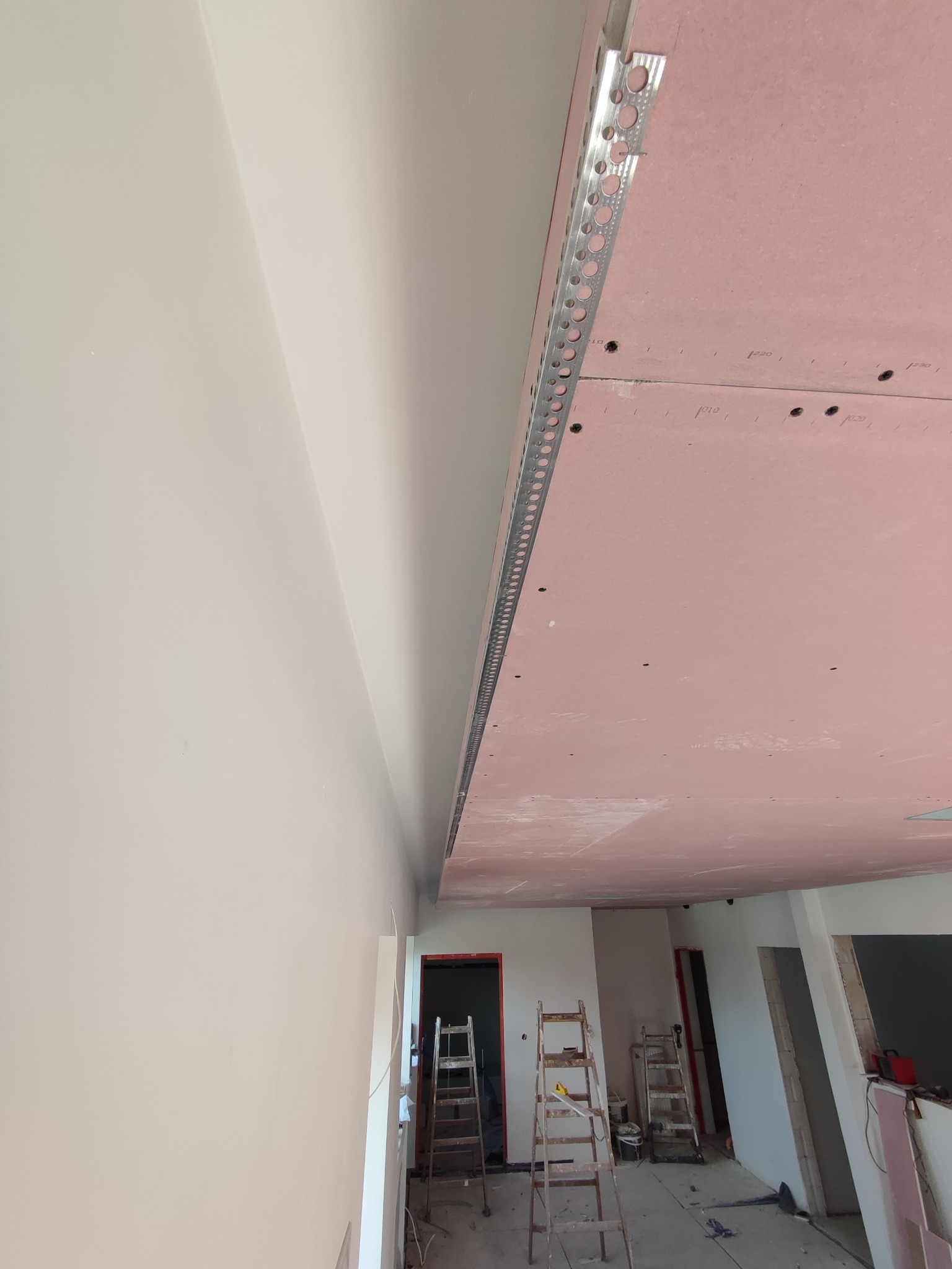 Montaż płyty gipsowo-kartonowej na suficie z widoczną metalową listwą wykończeniową i drabiny w tle.