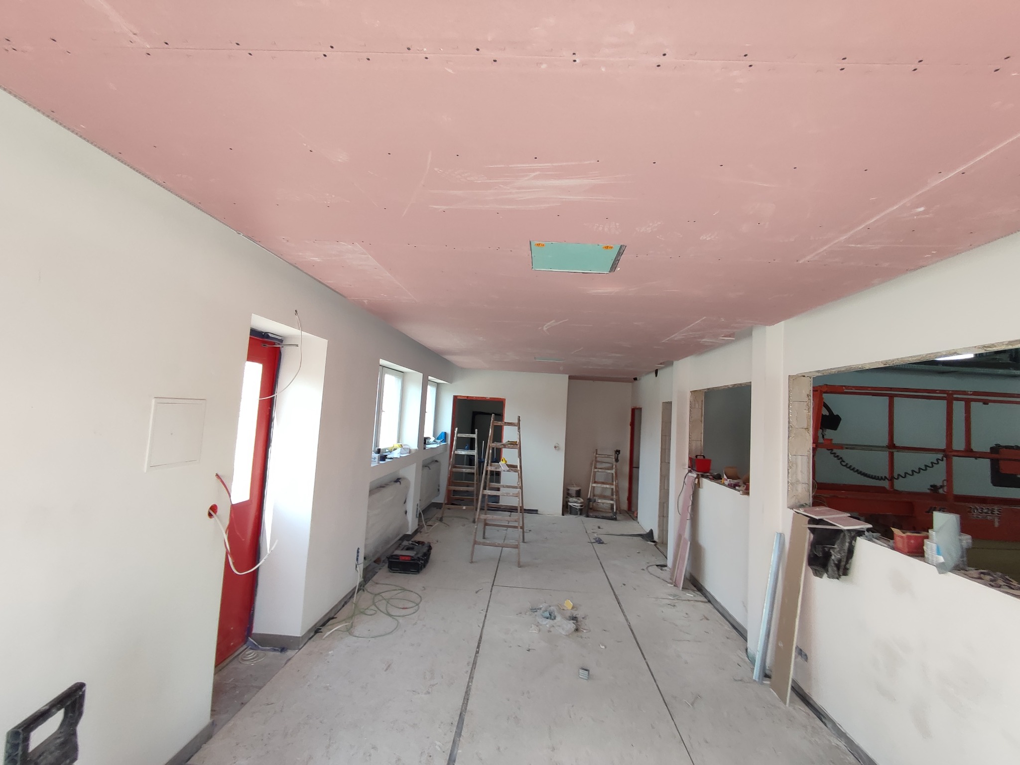 Wnętrze w trakcie remontu: różowe płyty na suficie, widoczne łączenia, ściany przygotowane do malowania, drabiny i narzędzia budowlane w pomieszczeniu.