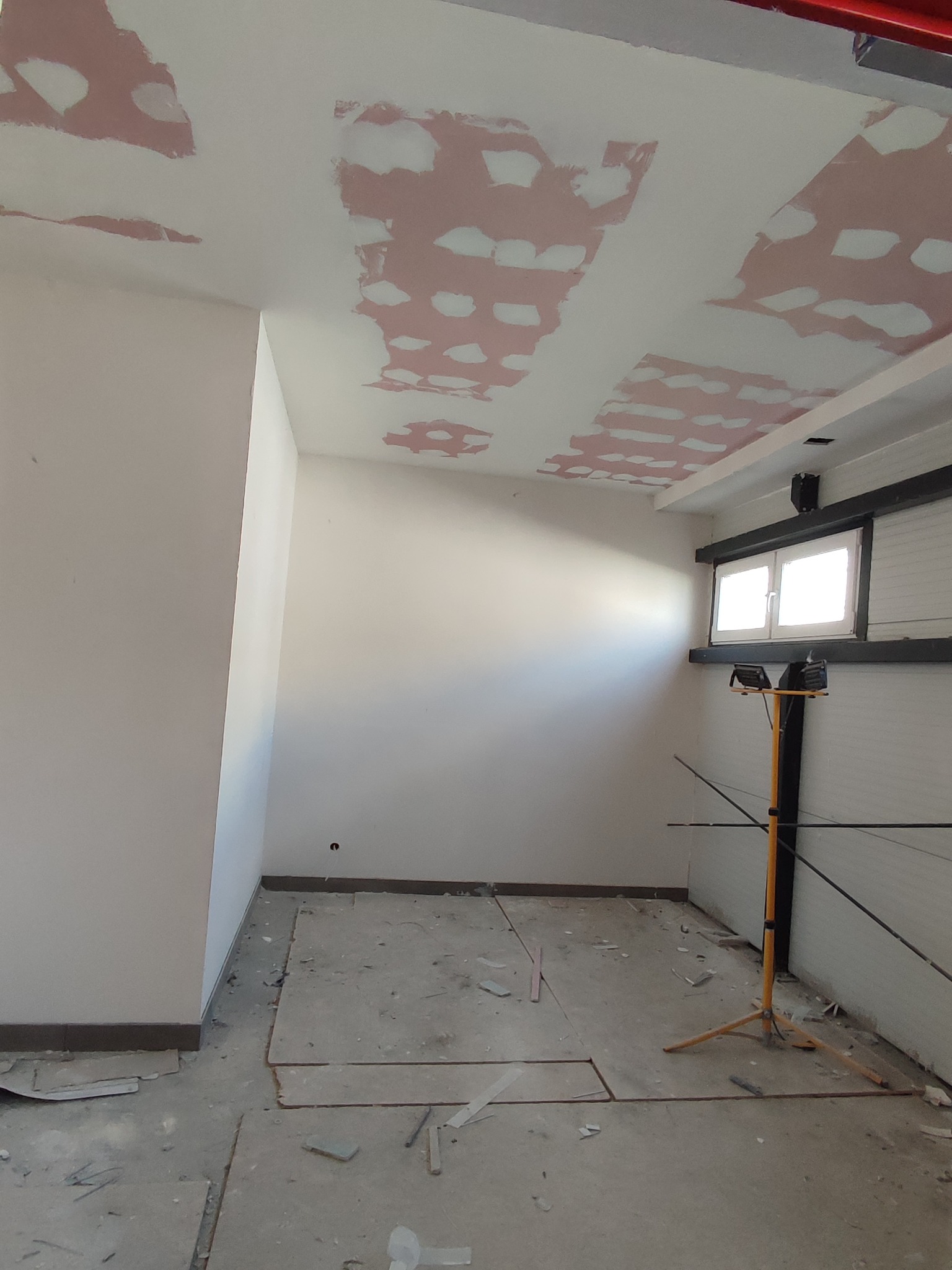 Wnętrze w trakcie remontu: sufit z widocznymi łatami szpachli, ściany przygotowane do malowania, podłoga z resztkami materiałów budowlanych, lampa budowlana na statywie.