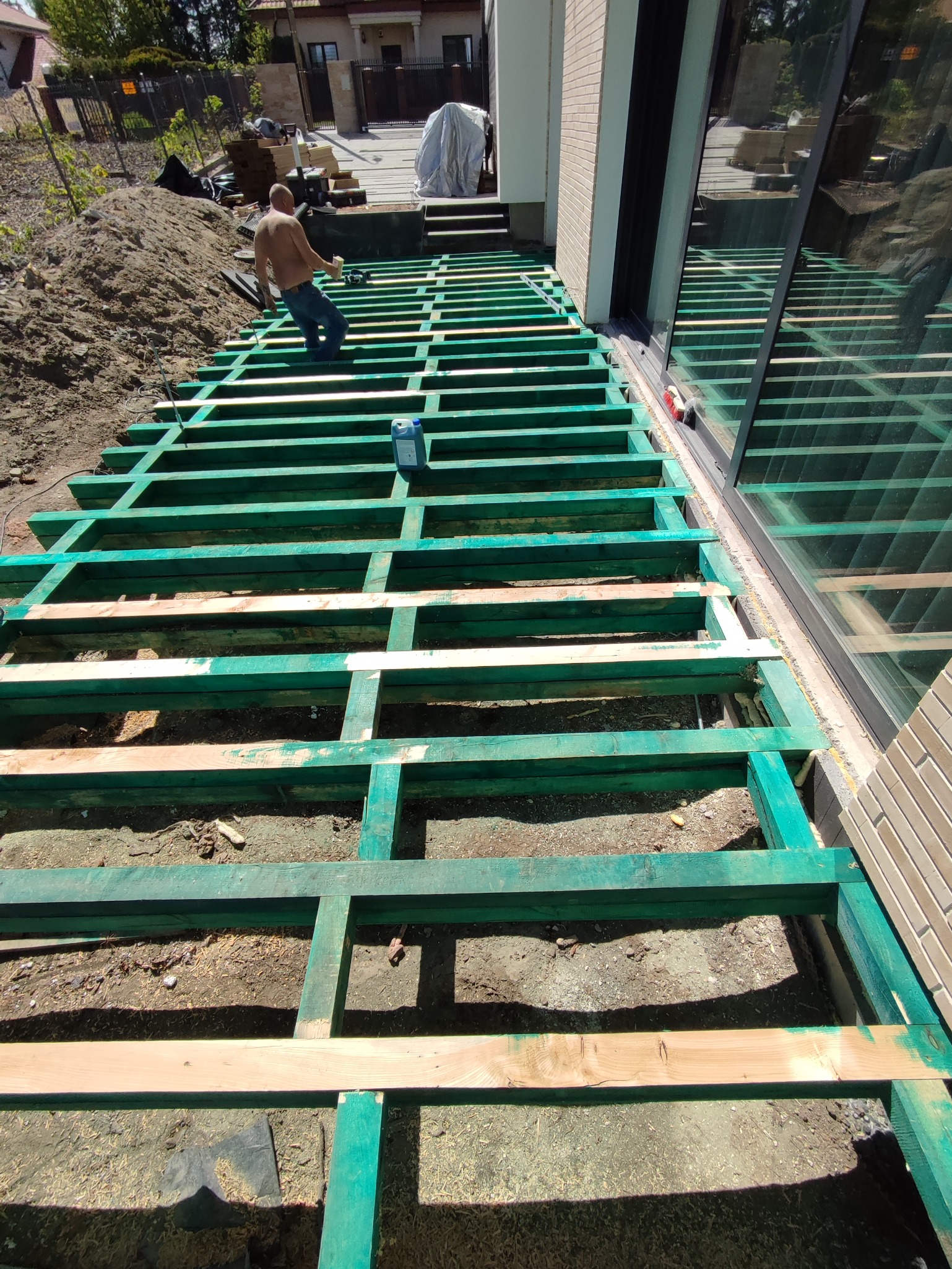 Konstrukcja nośna tarasu z impregnowanego drewna w trakcie budowy, widoczny pracownik bez koszulki i narzędzia, tło z domem w budowie.