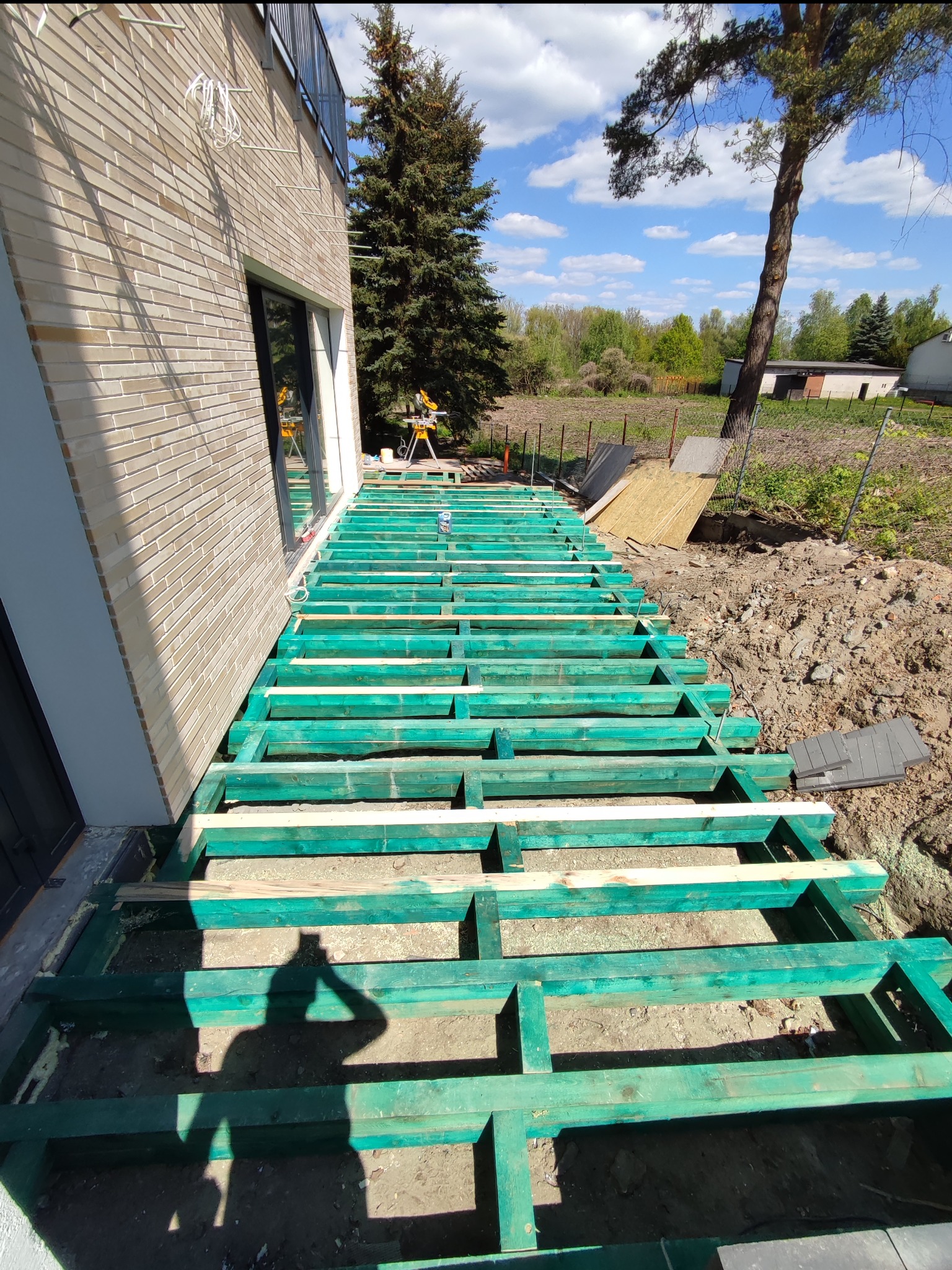 Konstrukcja nośna z impregnowanego drewna pod przyszły taras, widok z góry na szkielet, cień osoby fotografującej w lewym dolnym rogu, dom z cegły w tle, narzędzia na trawie.