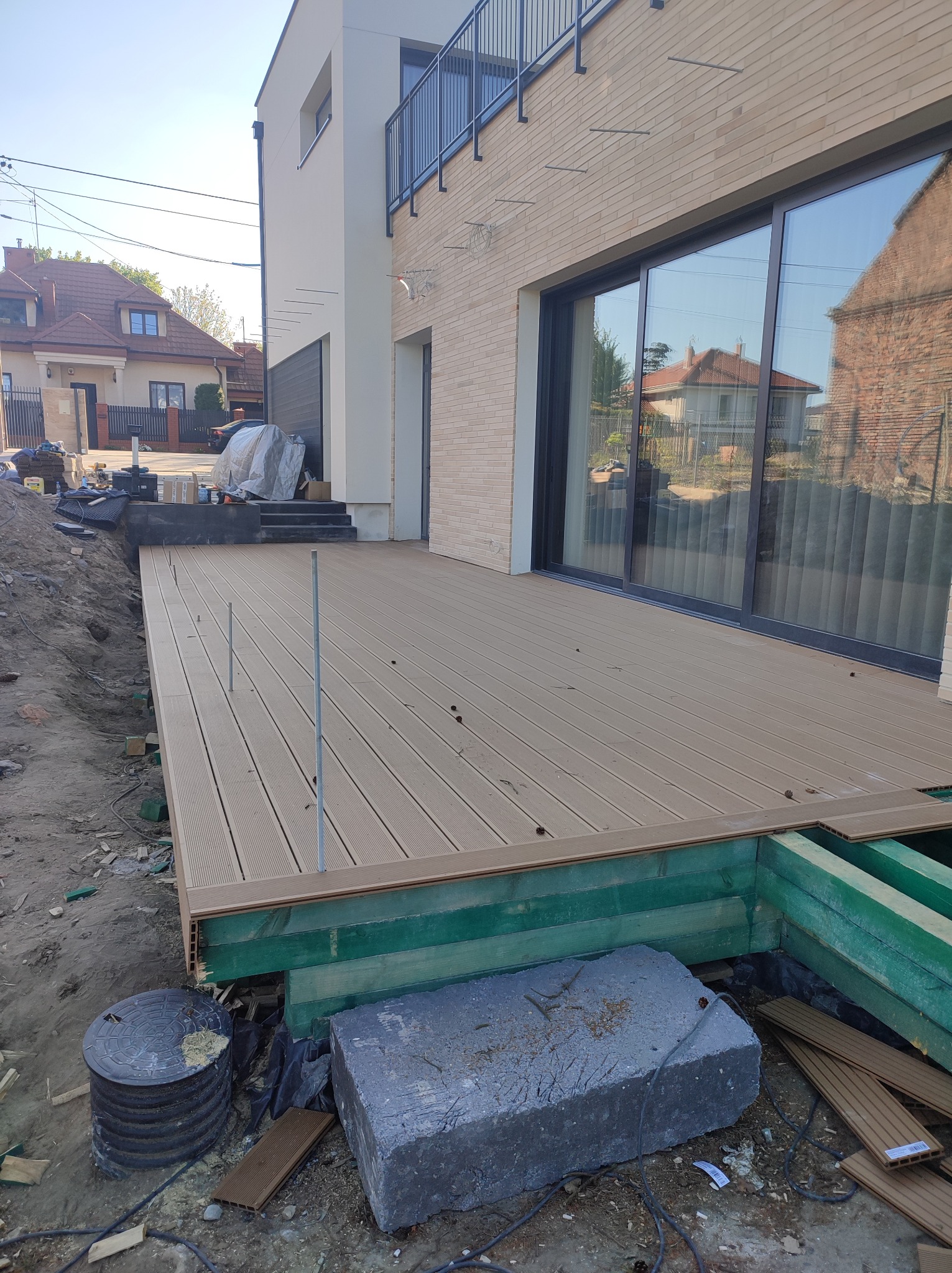 Montaż tarasu z desek kompozytowych przy nowoczesnym domu; widoczne elementy konstrukcyjne, podłoże z zielonej impregnacji oraz wystające metalowe pręty.