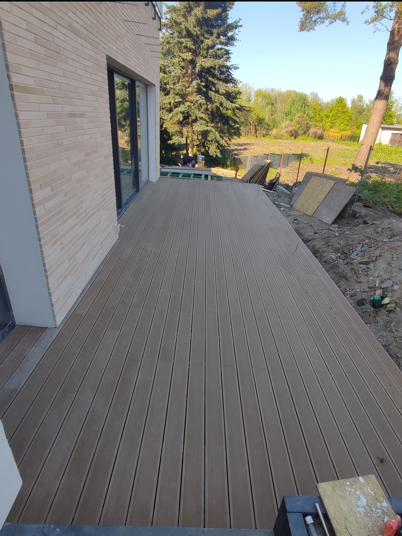 Montaż tarasu kompozytowego przy domu z jasnej cegły, widoczne deski w trakcie układania, przygotowane podłoże i narzędzia.