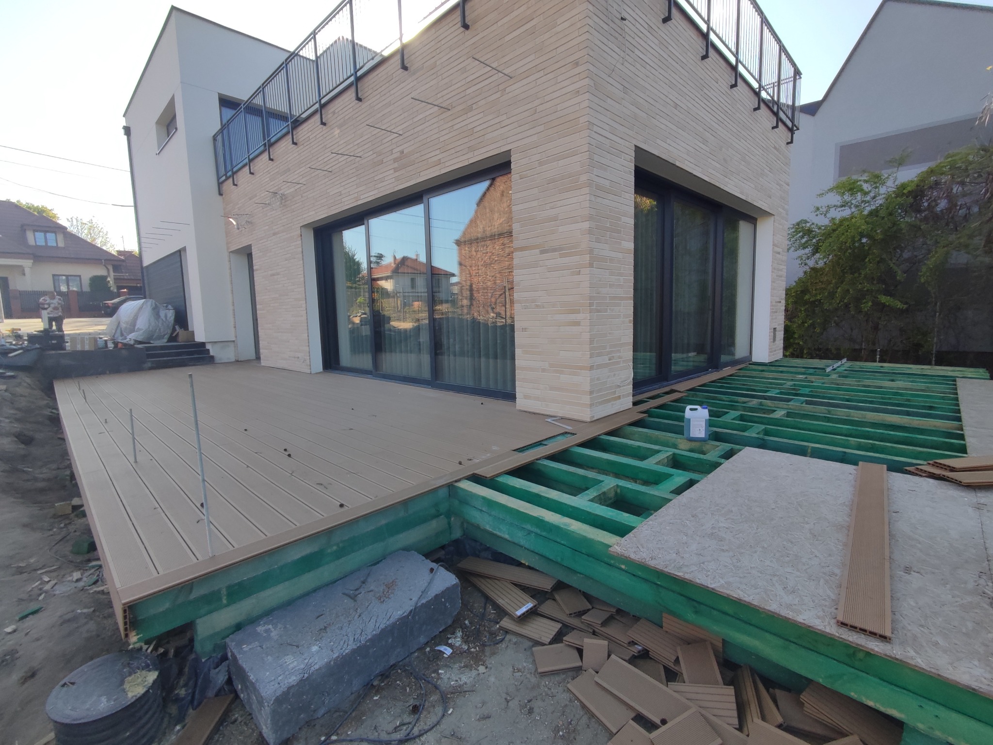 Montaż desek na tarasie z kompozytu na tle nowoczesnego domu z dużymi oknami i balkonem z czarną balustradą.