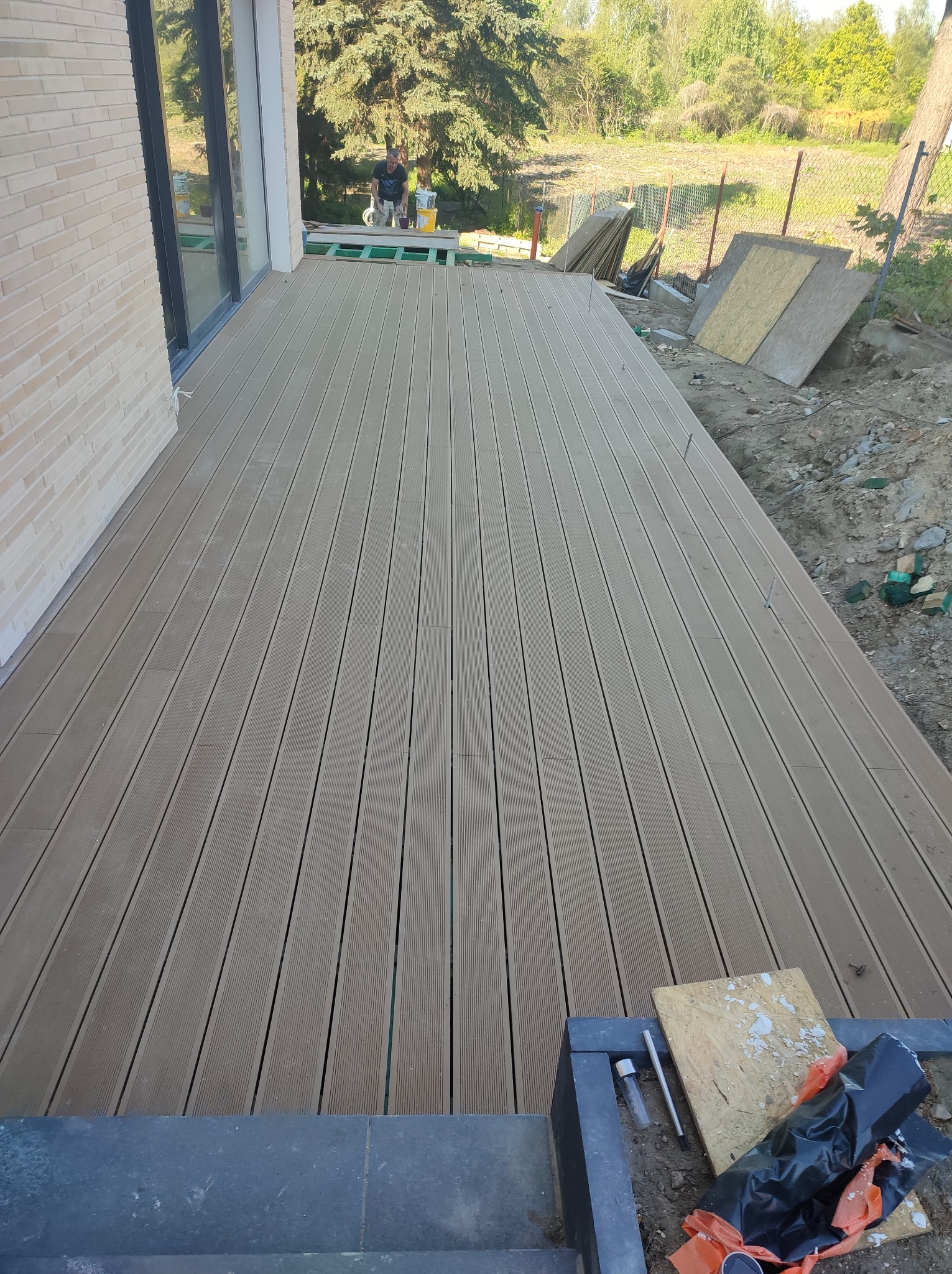 Nowo wybudowany taras z desek kompozytowych w kolorze jasnego brązu, widoczne elementy montażowe, w tle częściowo wykończona elewacja budynku i ogrodzenie.