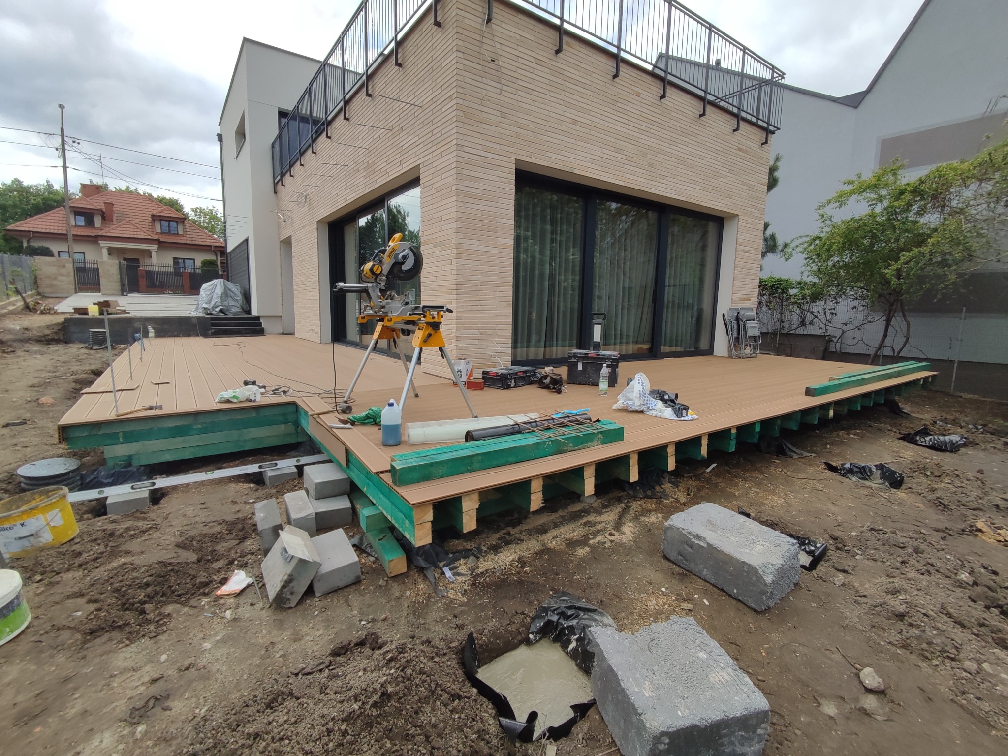Montaż tarasu z desek, widoczne podkonstrukcje na betonowych bloczkach, piła ukośnica na placu budowy, nowoczesny dom w tle.