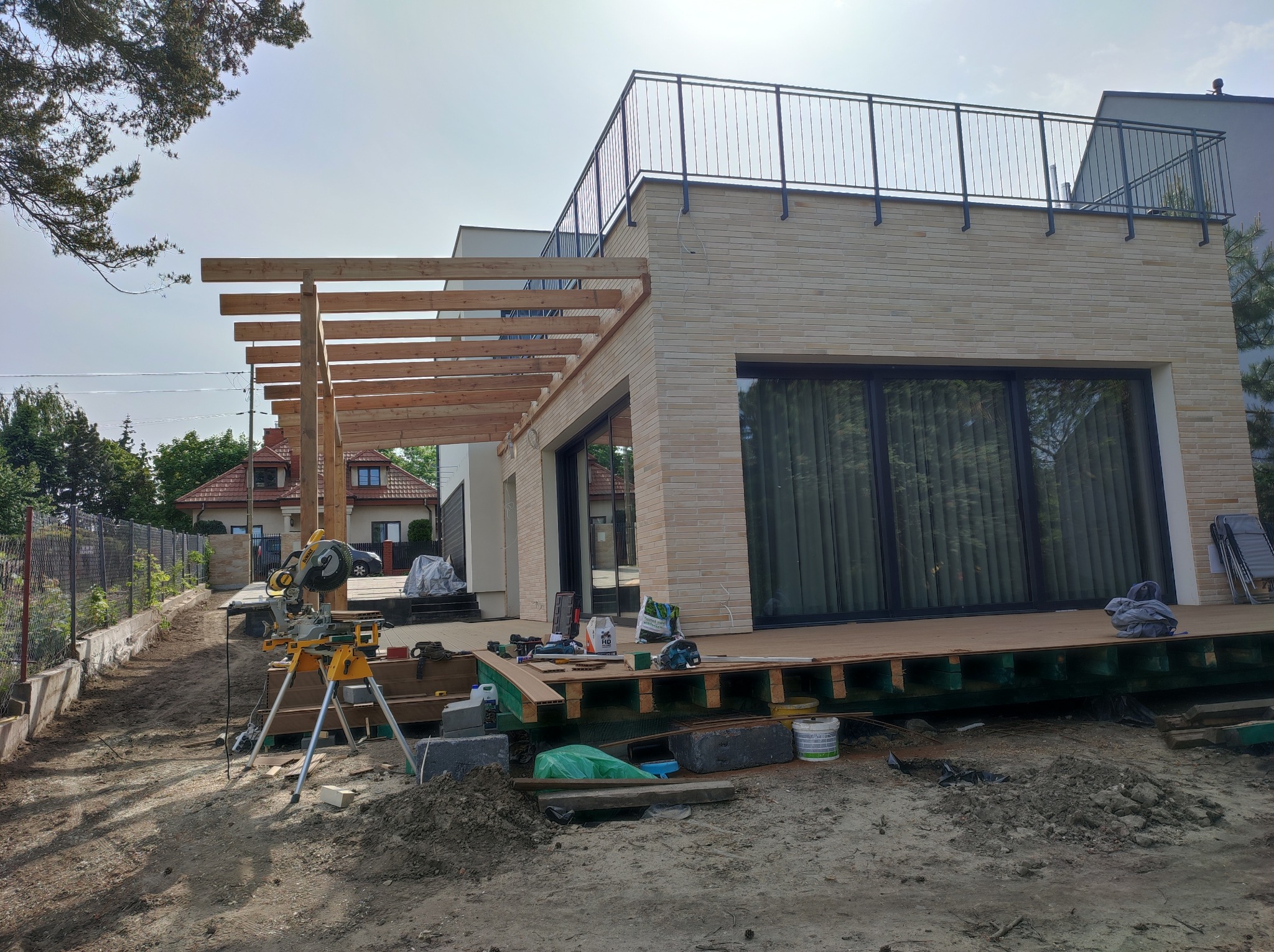 Budowa tarasu przy nowoczesnym domu z drewnianą konstrukcją pergoli i widoczną deską tarasową, narzędzia pracy w tle.