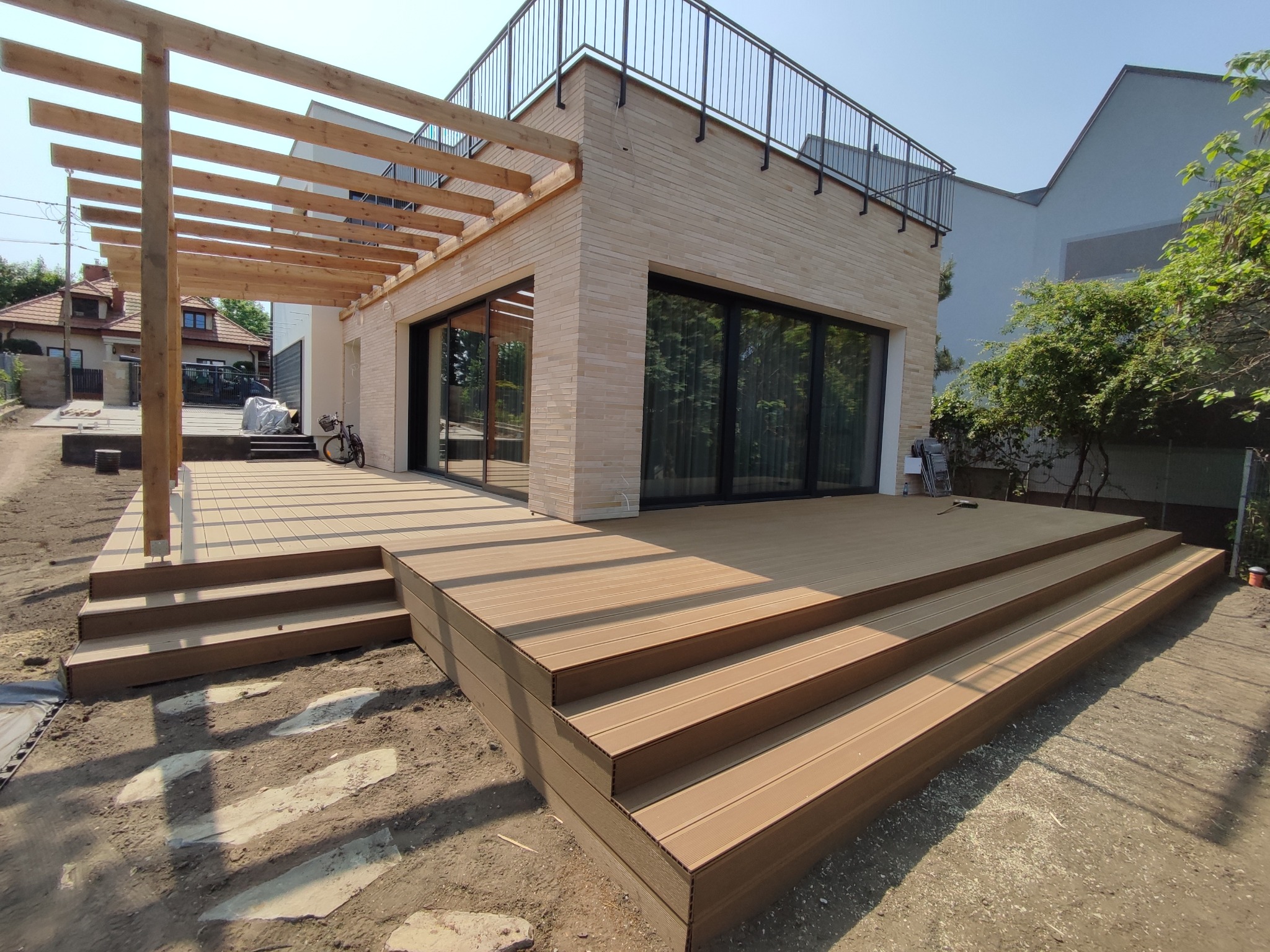 Nowoczesny dom z beżową elewacją i tarasem z desek kompozytowych, schody prowadzą na taras, pergola z drewnianych belek nad tarasem, rower oparty o ścianę budynku.