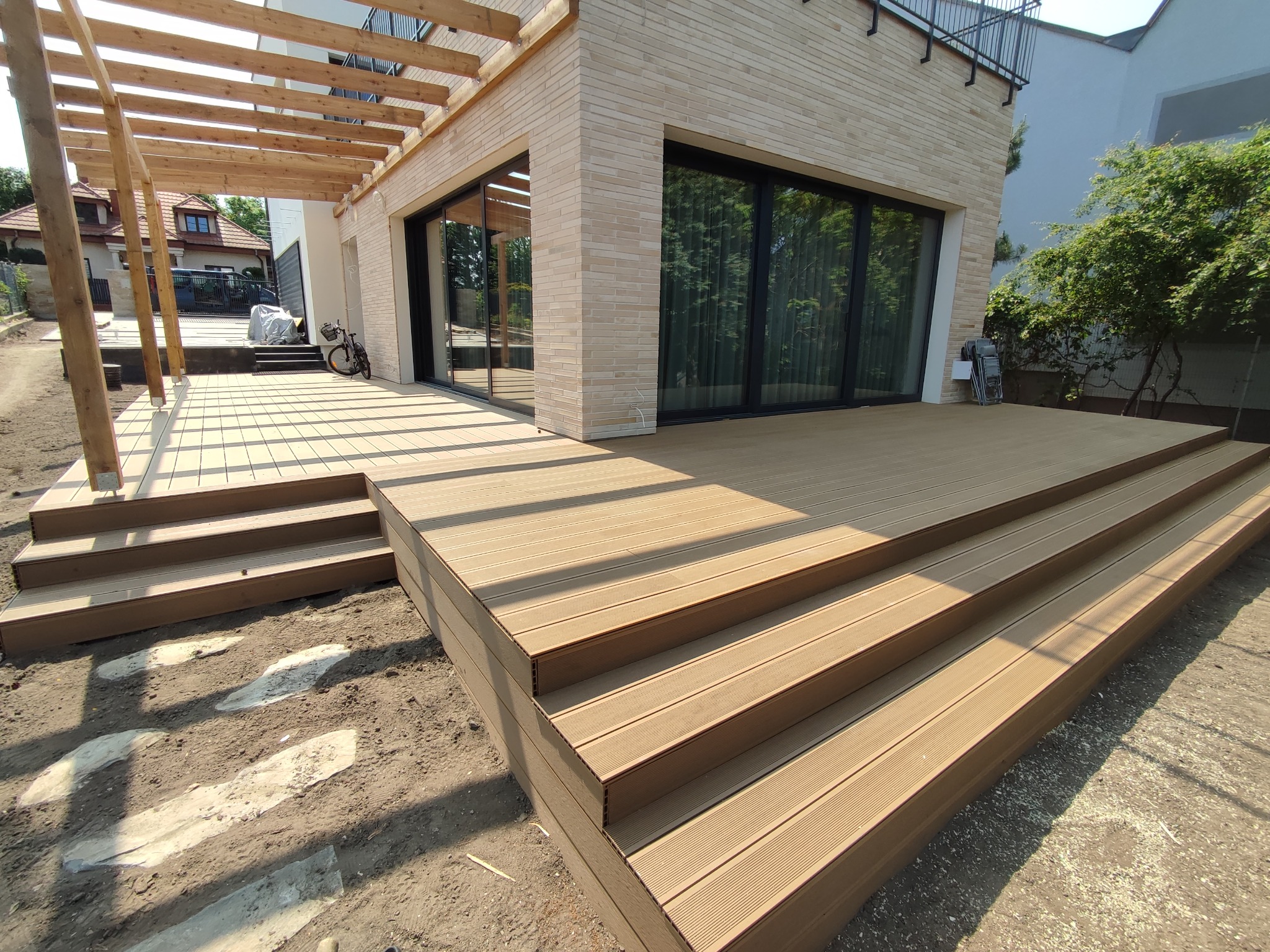 Nowoczesny taras z desek kompozytowych z czterostopniowymi schodami, przylegający do domu z beżowej cegły, z drewnianą pergolą nad częścią tarasu. Widoczne ślady montażu i kamienne płyty w ziemi.