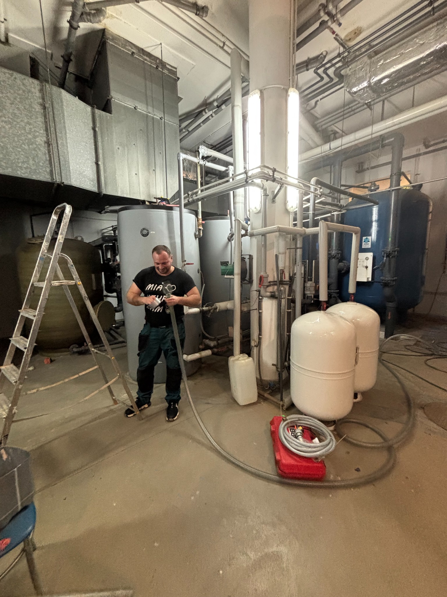 Instalator łączy rury w kotłowni. Widoczne zbiorniki, rury, drabina i narzędzia. W tle skomplikowana instalacja rur pod sufitem.