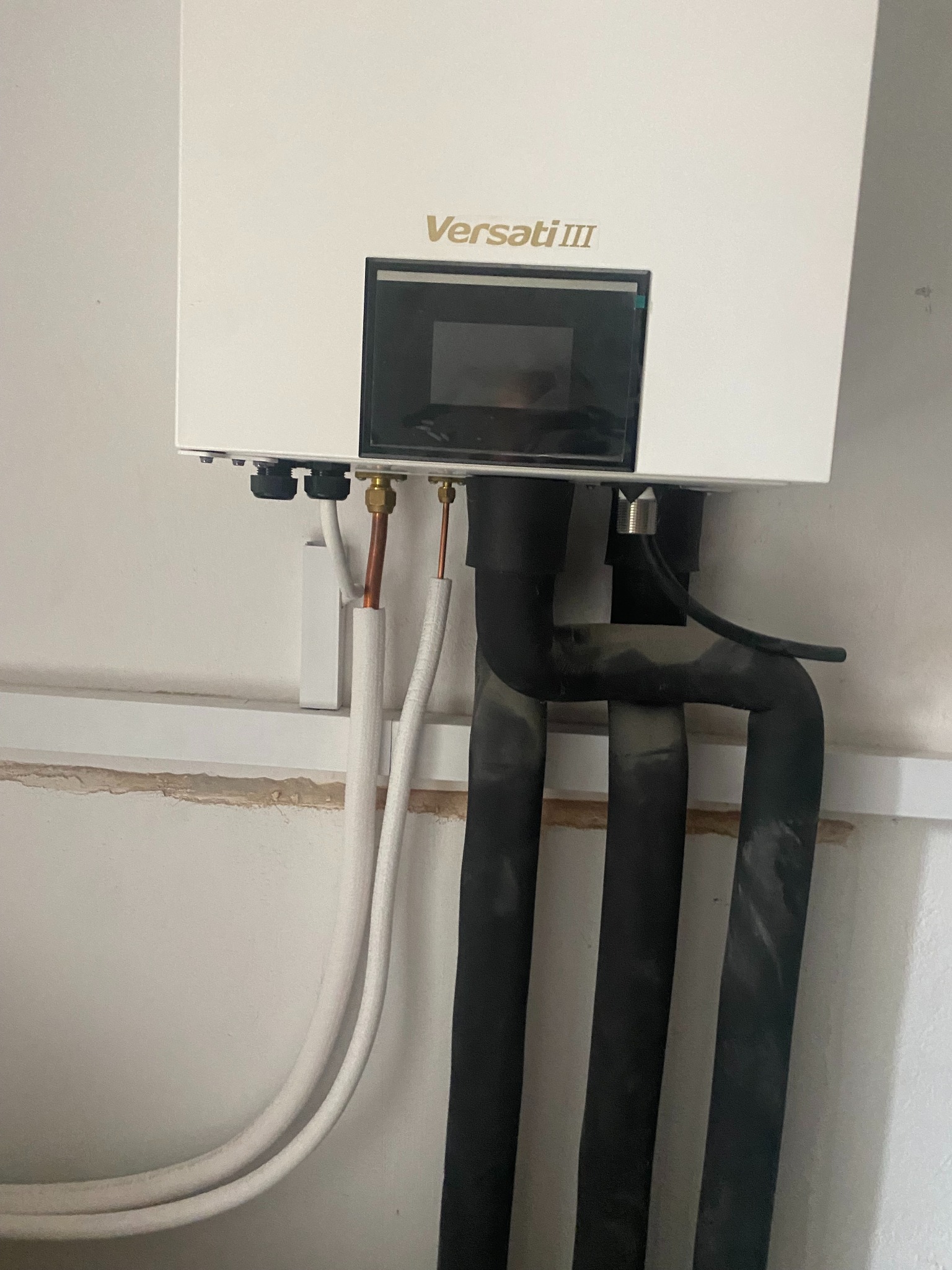 Biała pompa ciepła Versati III zamontowana na ścianie, z widocznymi rurami w izolacji, miedzianymi elementami i czarnymi przewodami. Ujęcie z bliska, instalacja w trakcie.