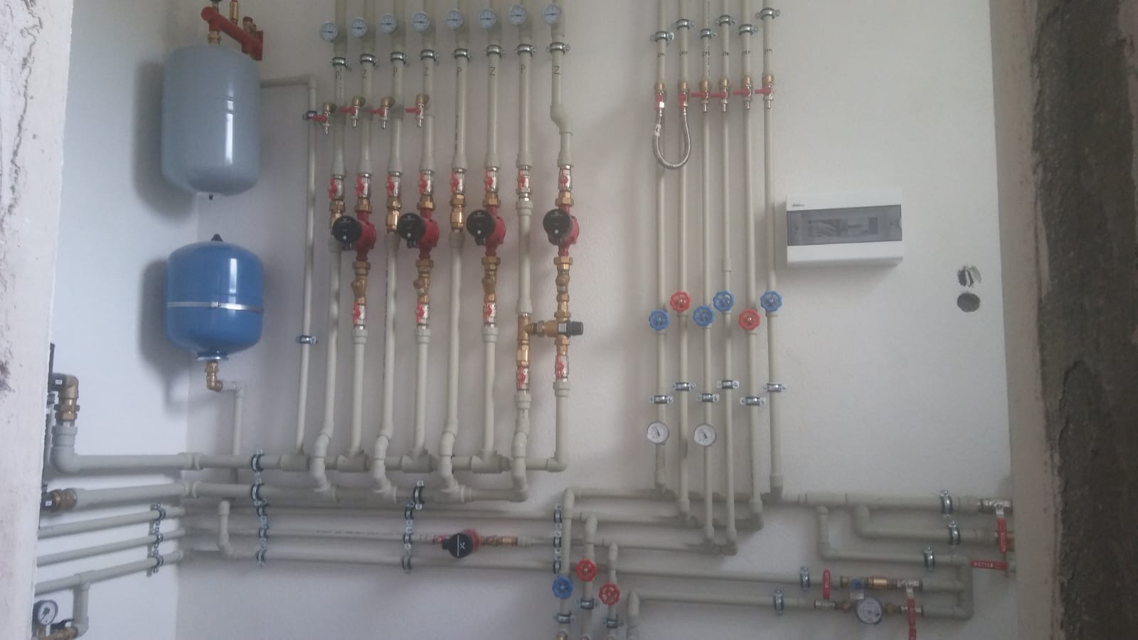 Rozbudowana instalacja hydrauliczna z licznymi rurami, zaworami i manometrami na białej ścianie w surowym wnętrzu. Widoczne zbiorniki wyrównawcze i rozdzielnica elektryczna.