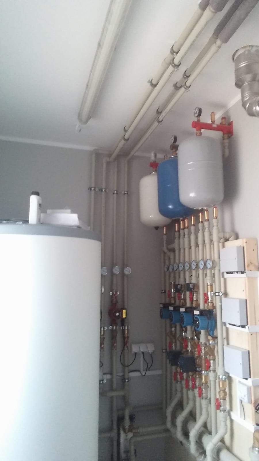 Biały zasobnik ciepłej wody użytkowej z niebieskim zbiornikiem wyrównawczym i licznymi rurami z zaworami w kotłowni. Instalacja grzewcza w pomieszczeniu technicznym.