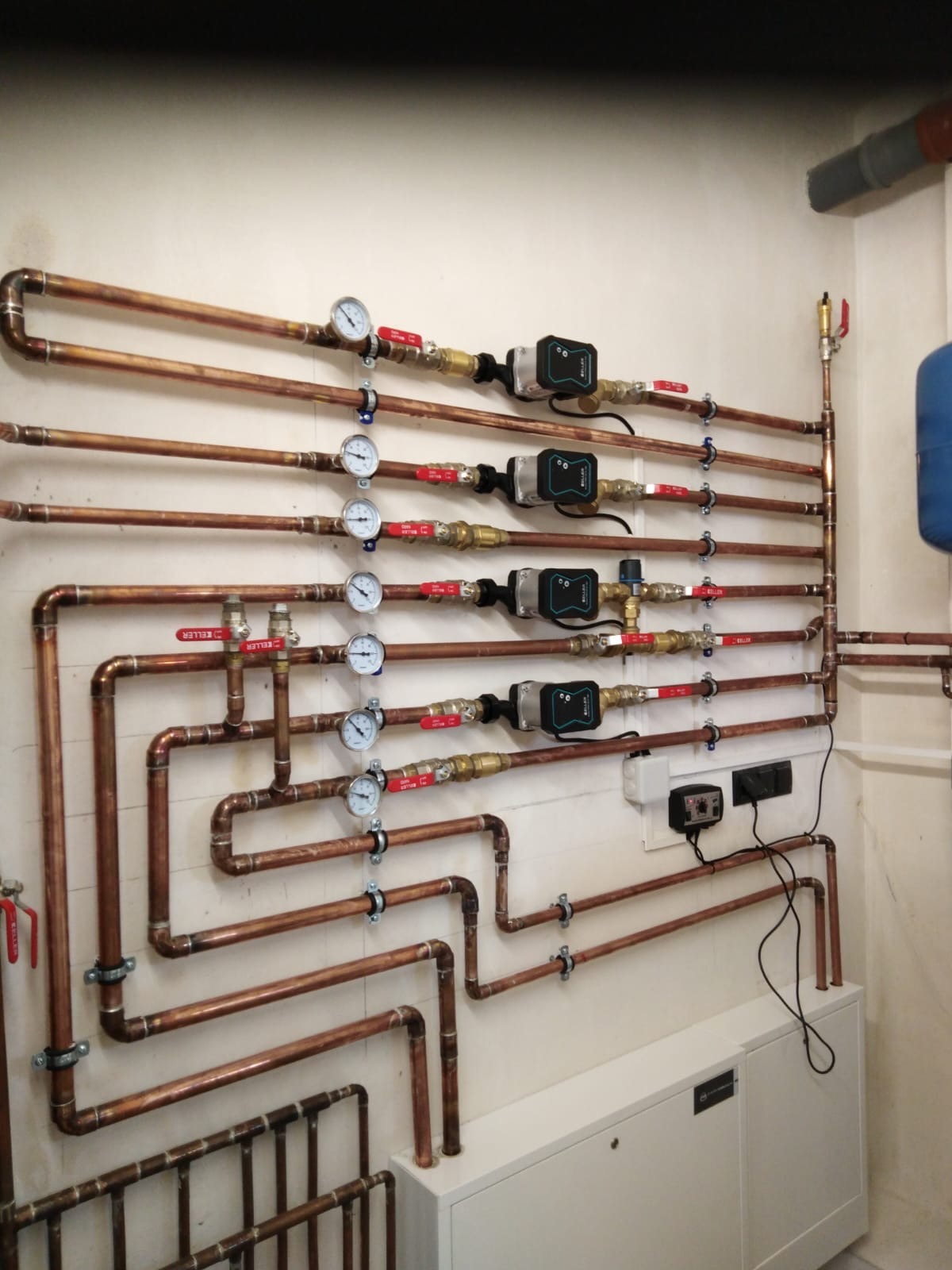 Miedziana instalacja hydrauliczna z manometrami, zaworami i pompami obiegowymi na białej ścianie. Widoczne elementy sterujące i szafka instalacyjna.
