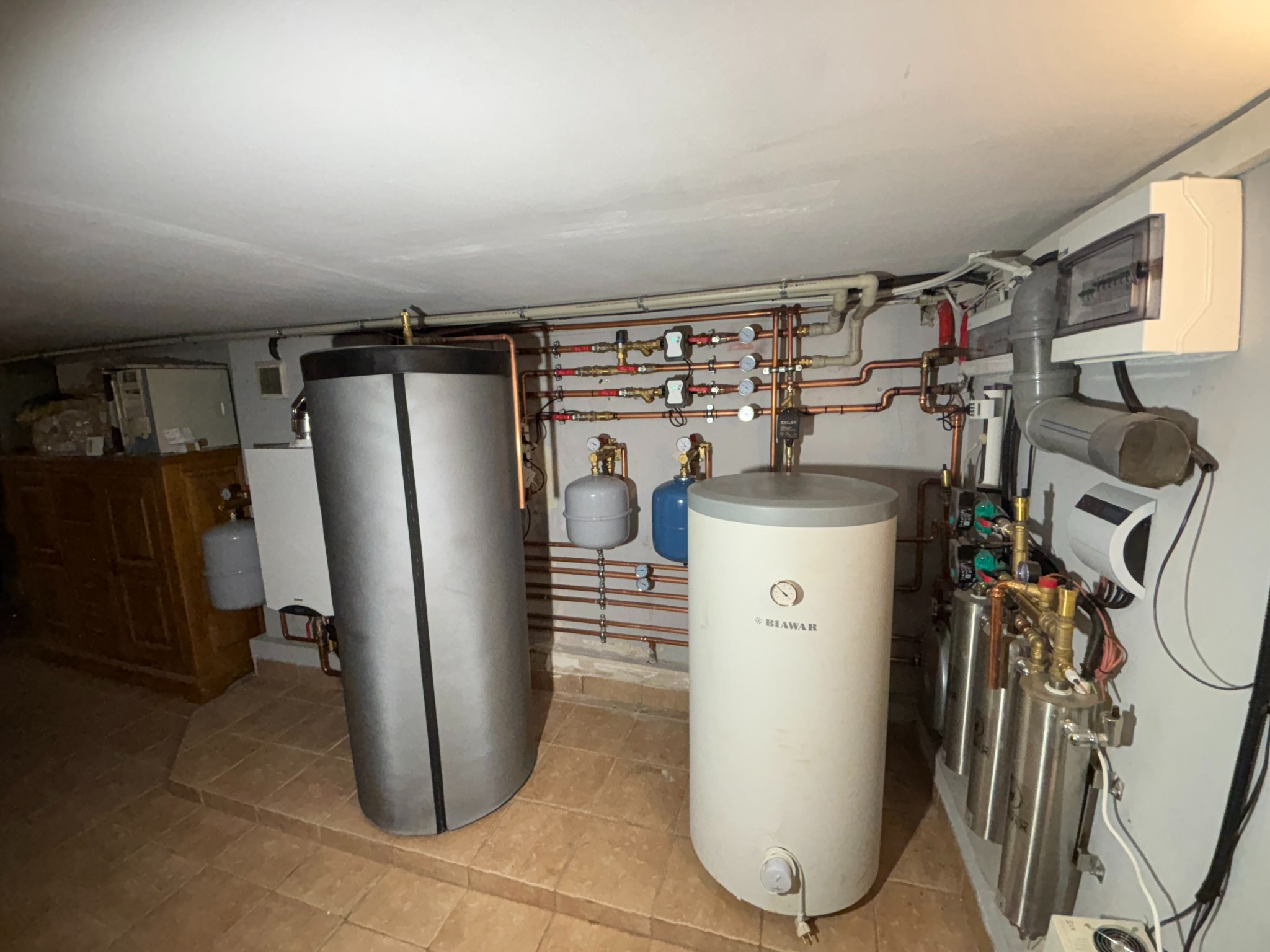 Złożona instalacja grzewcza w piwnicy: dwa zbiorniki, rury miedziane, zawory, manometry i rozdzielnica elektryczna. Biała ściana i brązowa podłoga z płytek.