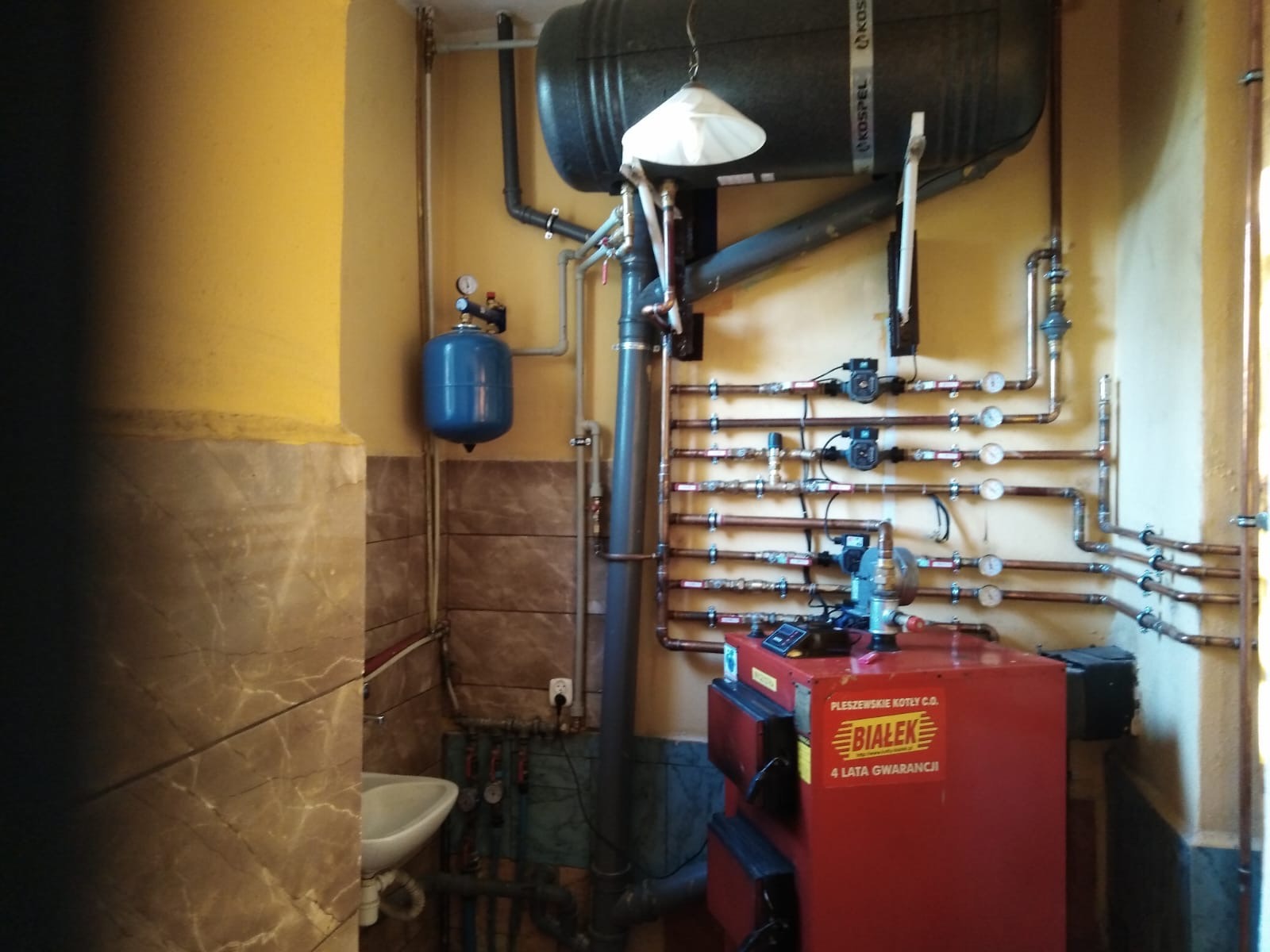 Czerwony piec CO 'Białek' z rozbudowaną instalacją hydrauliczną z miedzianych rur, zbiornikiem wyrównawczym i manometrami w kotłowni o żółtych ścianach.