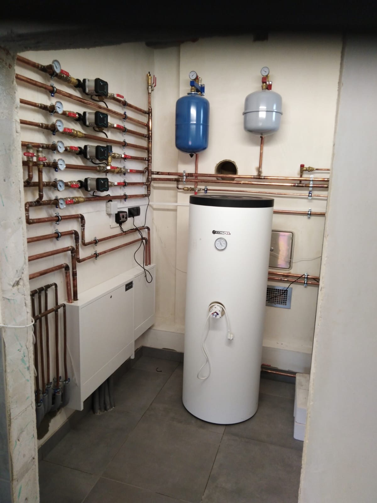 Biały zasobnik ciepłej wody Kospel z miedzianą instalacją, manometrami i zbiornikami wyrównawczymi w kotłowni. Widoczne liczne rury i zawory.