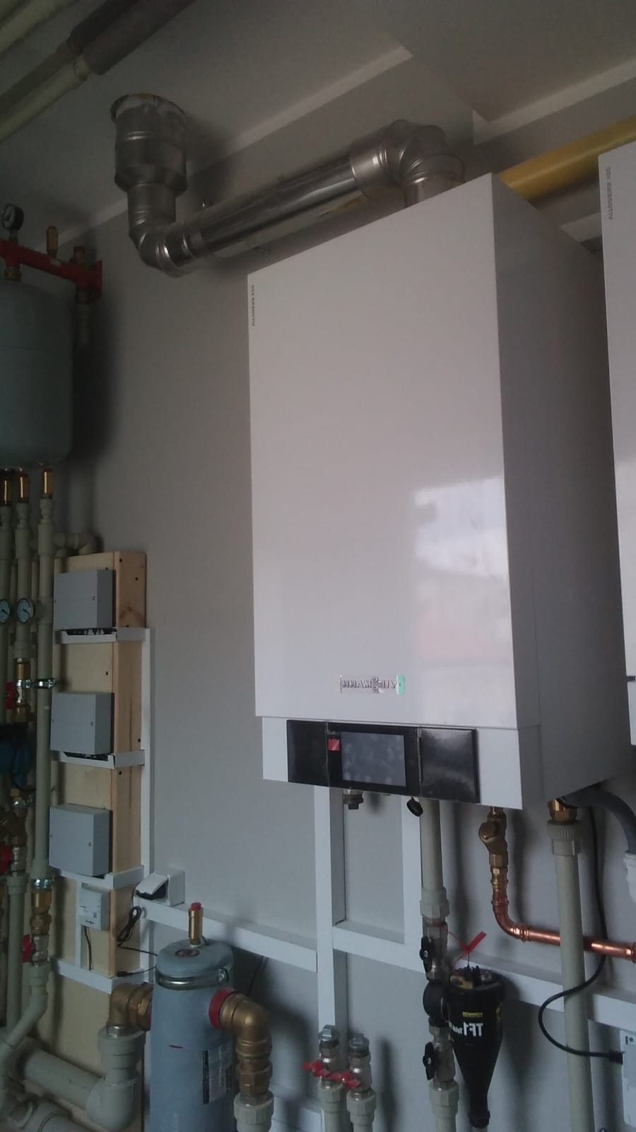 Biały, nowoczesny piec gazowy Viessmann z podłączeniami, rurami i zbiornikiem wyrównawczym na tle szarej ściany. Widoczne elementy instalacji CO i CWU.