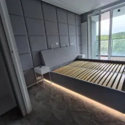 Szare łóżko z tapicerowanym wezgłowiem w panelach, podświetleniem LED i szafkami nocnymi na tle okna balkonowego.