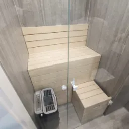 Wnętrze sauny z jasnymi drewnianymi ławkami ułożonymi w kilku poziomach, widoczny piec z kamieniami oraz szklane drzwi kabiny.