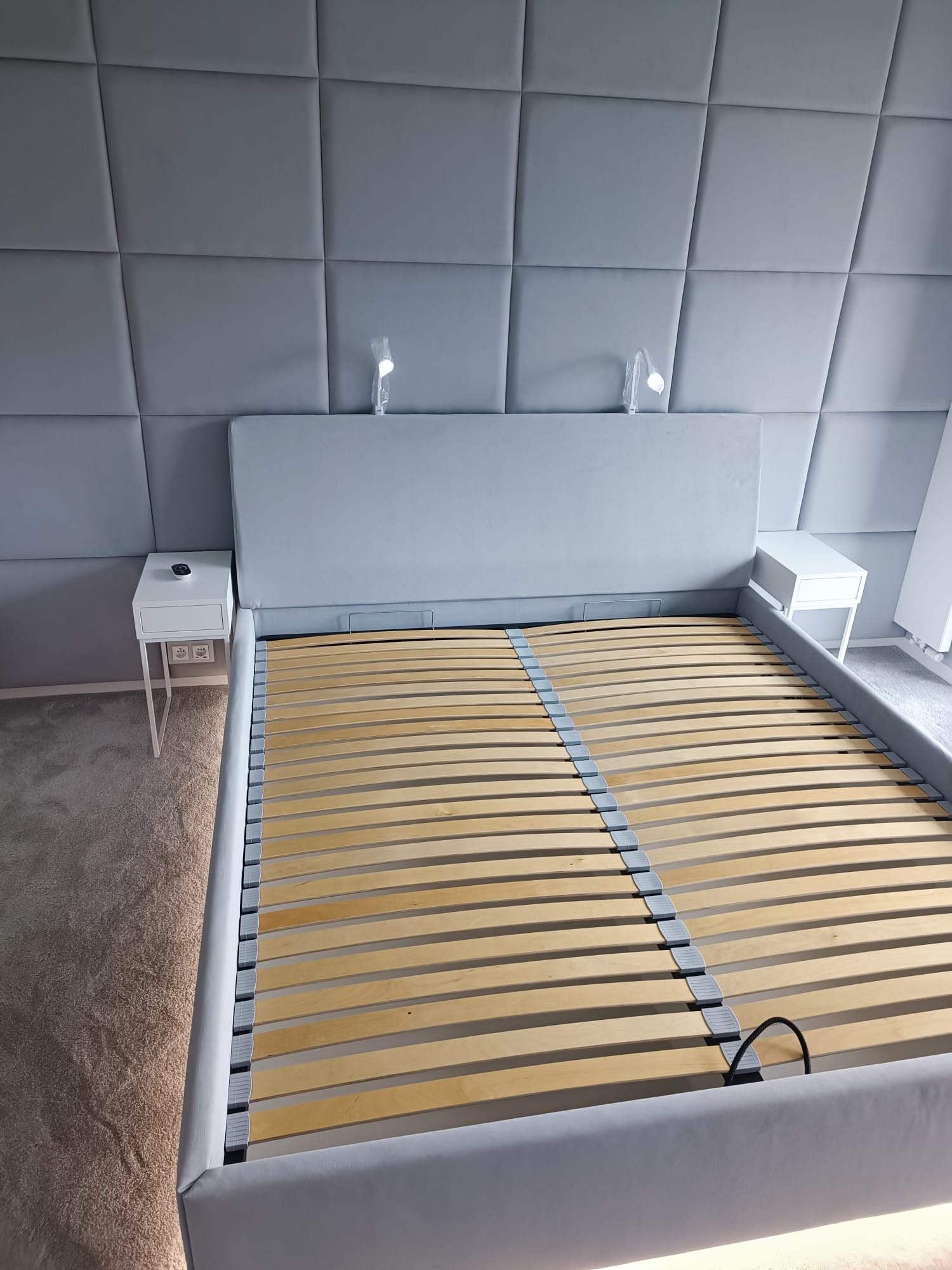 Szare łóżko z drewnianym stelażem i tapicerowanym zagłówkiem, dopasowane szafki nocne z lampkami, na tle ściany z tapicerowanymi panelami.