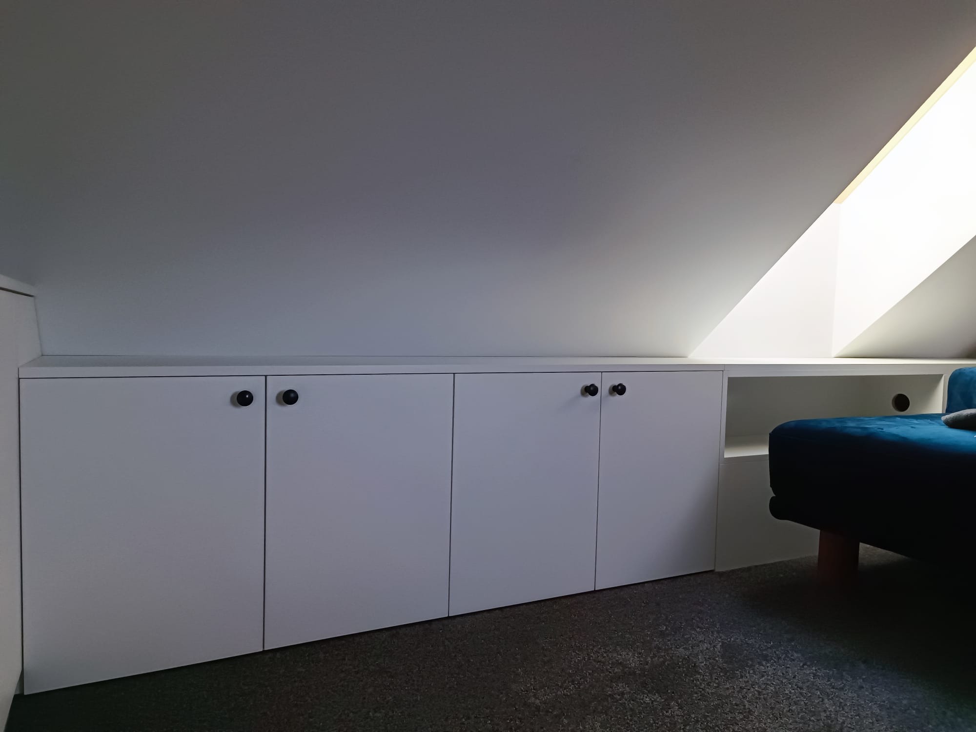 Biała zabudowa meblowa pod skosem dachu z czarnymi okrągłymi uchwytami, obok widoczny fragment niebieskiej sofy.