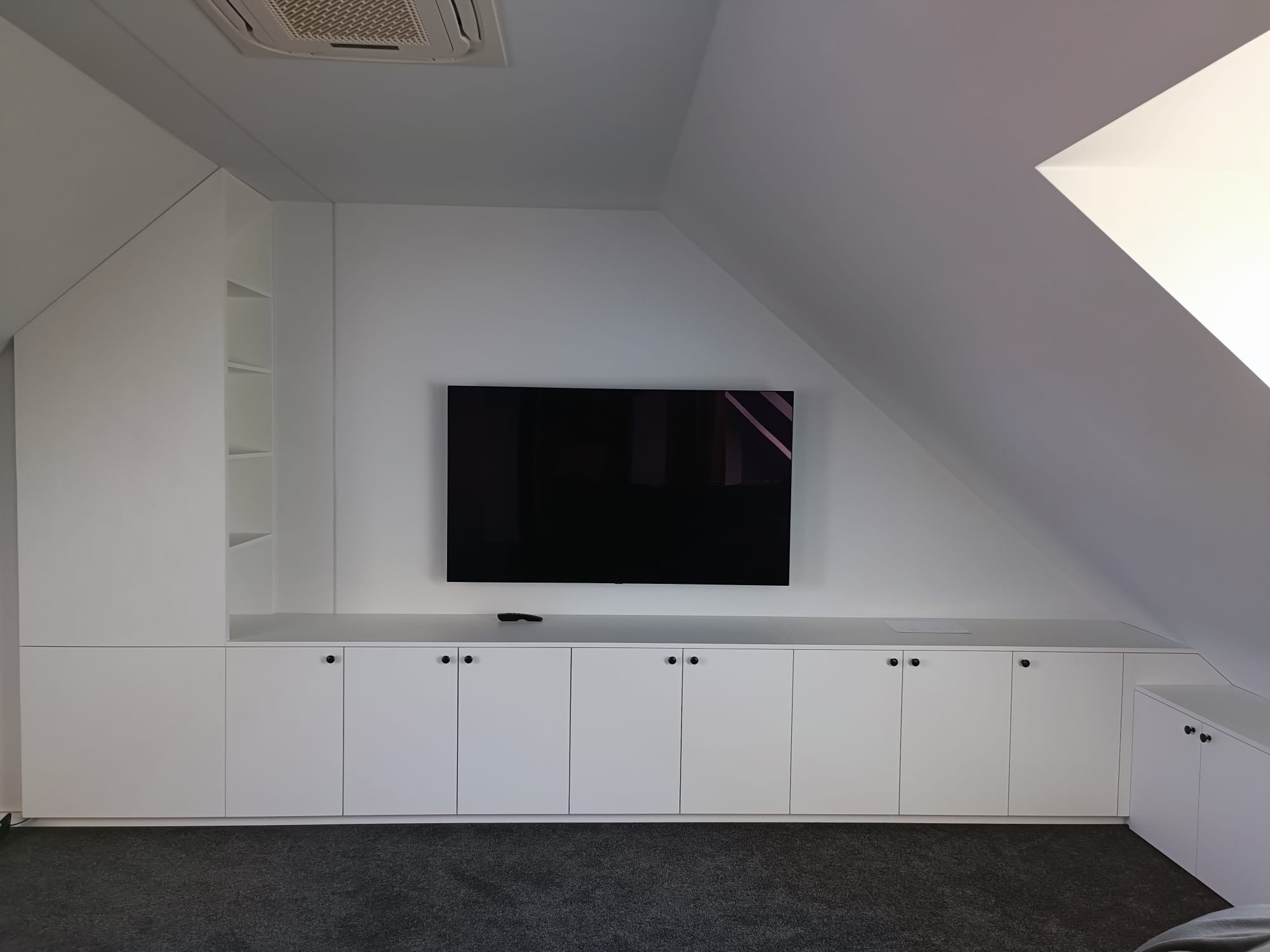 Biała zabudowa meblowa pod skosem na poddaszu, z szafkami z czarnymi okrągłymi uchwytami, półkami i miejscem na telewizor, klimatyzacja na suficie.