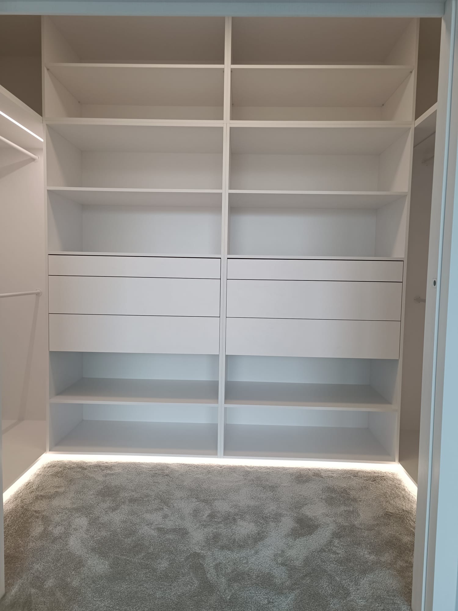 Wnętrze przestronnej, białej garderoby z półkami i szufladami, podświetlonej od dołu, z szarym dywanem na podłodze.