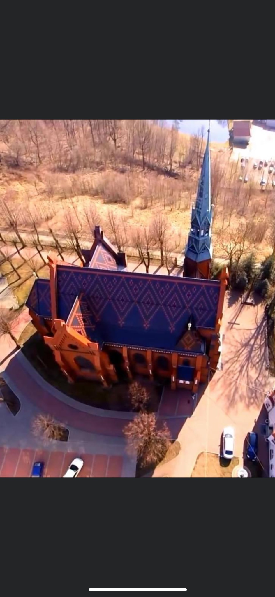 Widok z lotu ptaka na ceglany kościół z wysoką wieżą i wzorzystym, ciemnoniebieskim dachem, otoczony drzewami i parkingiem z samochodami.