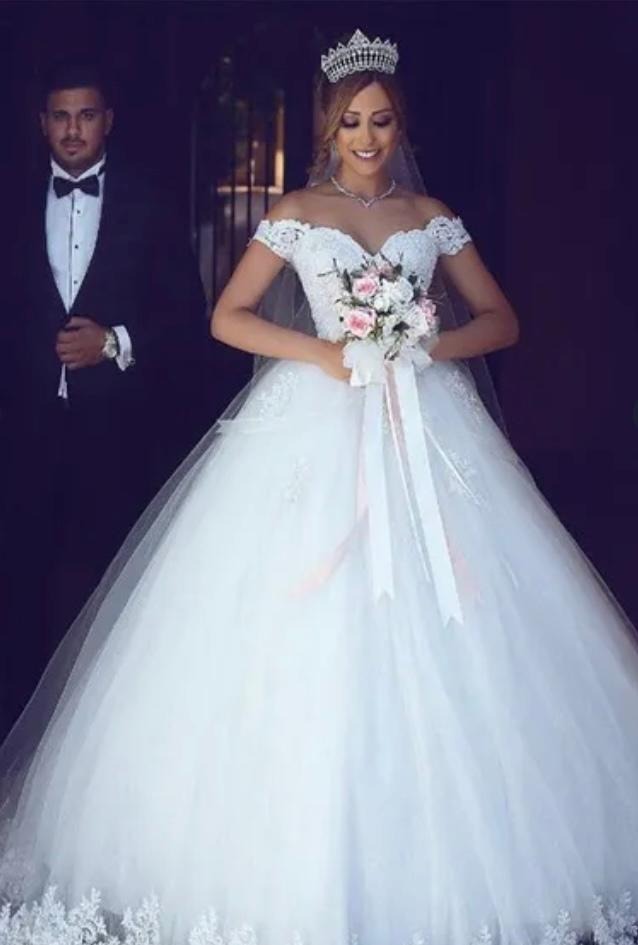 Panna młoda w koronie i sukni balowej z odkrytymi ramionami trzyma bukiet różowych i białych kwiatów, obok mężczyzna w smokingu z muchą.